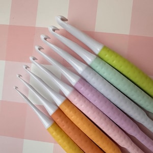 May include: A selection of crochet hooks featuring white metal hooks and vibrantly coloured, textured handles. The handles are in shades of yellow, orange, pink, purple, light blue, and green. The hooks are arranged diagonally against a pink and white checkered background.