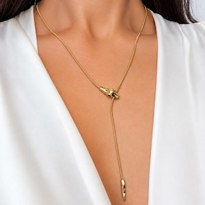 May include: A gold-toned lariat necklace featuring a thin, flexible snake chain. The necklace includes a smooth, organic-shaped metal accent piece that slides along the chain, ending in a slender, tapered metal drop at the bottom of the strand.
