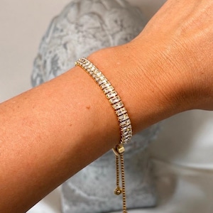 Op de afbeelding: Een verstelbare, goudkleurige armband met een doorlopende rij heldere, rechthoekige baguette-geslepen stenen. De armband heeft een schuifsluiting met twee dunne, hangende gouden kettinkjes, elk afgewerkt met een klein gouden kraaltje aan het uiteinde.