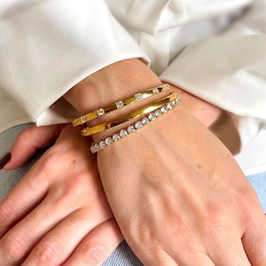 Op de afbeelding: Drie gouden armbanden met heldere zirkonia-stenen. De bovenste armband heeft een rij vierkante stenen, de middelste armband is een gladde band en de onderste armband heeft een rij ronde stenen.