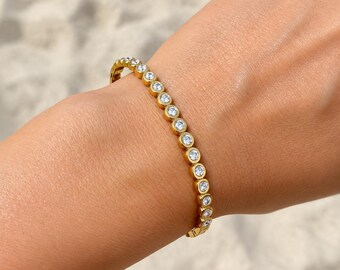 Puede incluir: Una pulsera de tenis en tono dorado que presenta una fila continua de piedras redondas y transparentes engastadas en bisel. La pulsera está construida con monturas metálicas circulares individuales que sujetan cada piedra, creando un diseño de cadena.