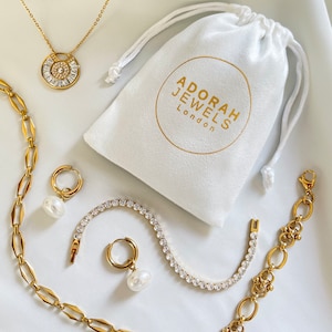 May include: Gold-toned jewelry set including a necklace, earrings, bracelet, and chain. The necklace features a circular pendant with a central stone and surrounding baguette-cut stones. A white drawstring bag with the text "ADORAH JEWELS London" is also present.