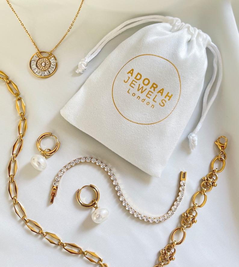 May include: Gold-toned jewelry set including a necklace, earrings, bracelet, and chain. The necklace features a circular pendant with a central stone and surrounding baguette-cut stones. A white drawstring bag with the text "ADORAH JEWELS London" is also present.