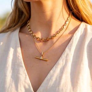 May include: A close-up shot of two gold necklaces. The top necklace has a chunky chain design, while the bottom necklace features a delicate chain with a T-bar pendant. The pendant has a small, clear gemstone. The necklaces are worn over a cream-colored top.