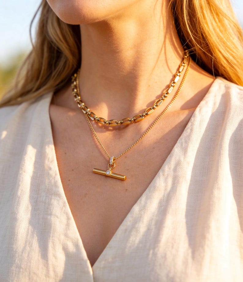 May include: A close-up shot of two gold necklaces. The top necklace has a chunky chain design, while the bottom necklace features a delicate chain with a T-bar pendant. The pendant has a small, clear gemstone. The necklaces are worn over a cream-colored top.