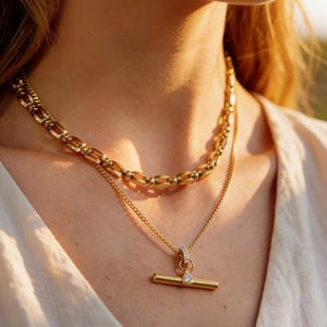 May include: A close-up shot of two gold necklaces. The top necklace is a chunky chain, while the bottom one features a delicate chain with a bar pendant and a small, clear gemstone. The jewelry is displayed on a person wearing a cream-colored top.