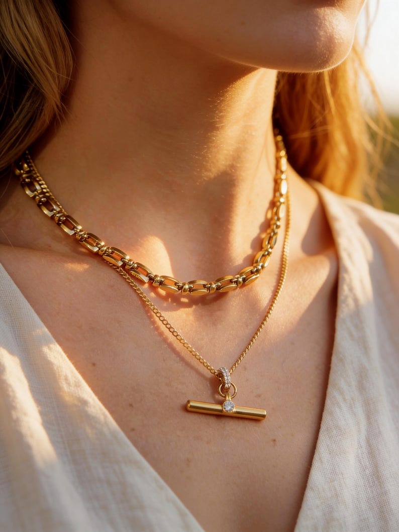May include: A close-up shot of two gold necklaces. The top necklace is a chunky chain, while the bottom one features a delicate chain with a bar pendant and a small, clear gemstone. The jewelry is displayed on a person wearing a cream-colored top.