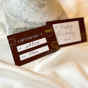 May include: Two gift cards on a white satin surface. One card is brown with gold accents and the text "A GIFT FOR YOU!" and "To: Alice" and "From: Tom". The other card has handwritten text that says "Happy Birthday".