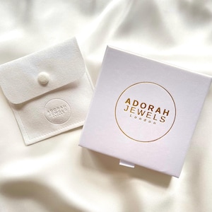 May include: A white jewelry box and pouch set. The box has a gold circular logo with the text "ADORAH JEWELS London". The pouch is a small, square, white fabric with a button closure and a circular logo that reads "ADORAH JEWELS".