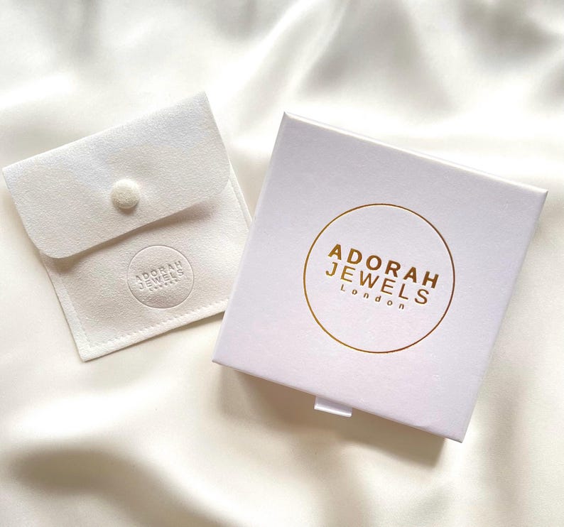 May include: A white jewelry box and pouch set. The box has a gold circular logo with the text "ADORAH JEWELS London". The pouch is a small, square, white fabric with a button closure and a circular logo that reads "ADORAH JEWELS".
