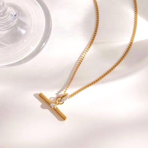 May include: A gold-colored necklace with a T-bar pendant. The pendant features a small, round, clear gemstone and is connected to a delicate chain. The necklace is displayed on a white surface.