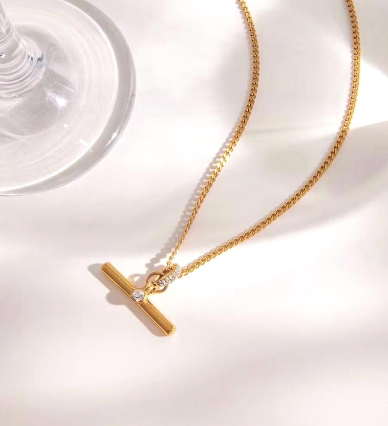 May include: A gold-colored necklace with a T-bar pendant. The pendant features a small, round, clear gemstone and is connected to a delicate chain. The necklace is displayed on a white surface.