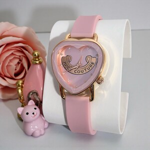 May include: A light pink analog watch with a heart-shaped gold-tone case and a matching pink silicone strap. The watch face features the text Juicy Couture in a gold banner, small crystal hour markers, and gold-tone hands on a pale pink background.