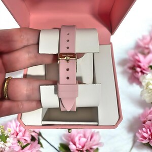 May include: A light pink silicone watch strap with a gold-tone metal buckle featuring the text Juicy Couture. The strap is secured around a white watch display cushion inside a light pink hinged presentation box with a smooth finish.