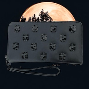 May include: Black wallet featuring embossed spider designs. The wallet has a wrist strap and a zip closure. The background shows a full moon and silhouetted trees, creating a spooky theme.