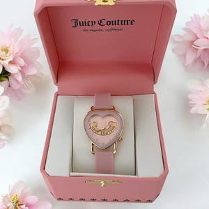 May include: A pink Juicy Couture watch with a heart-shaped face and rose gold accents, presented in a matching pink hinged box. The watch features a pink strap and the text Juicy Couture los angeles, california printed inside the box lid.