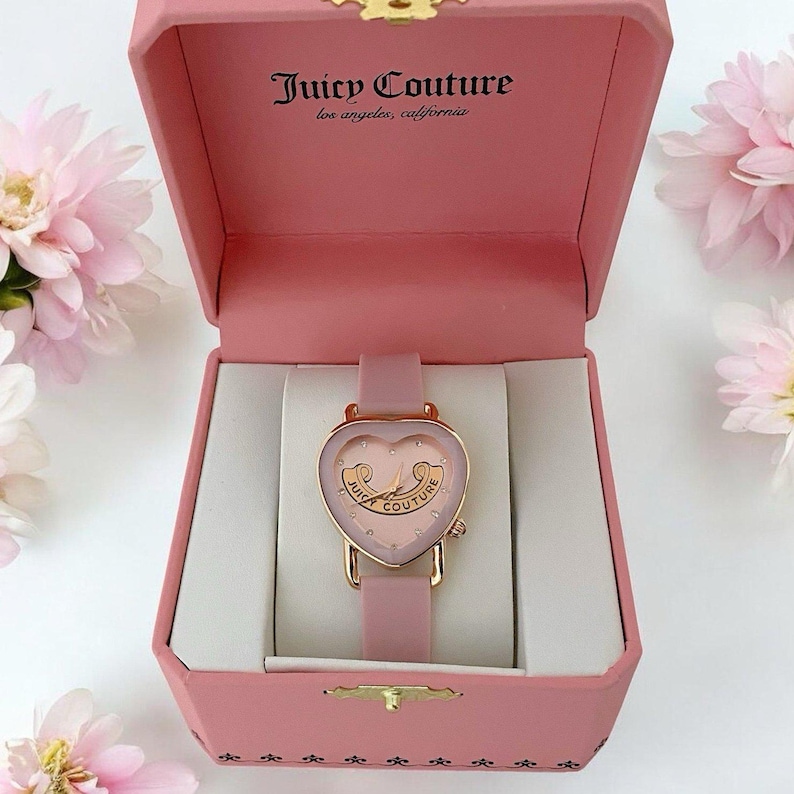 May include: A pink Juicy Couture watch with a heart-shaped face and rose gold accents, presented in a matching pink hinged box. The watch features a pink strap and the text Juicy Couture los angeles, california printed inside the box lid.