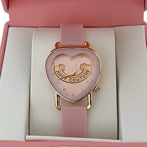 May include: A heart-shaped analog watch with a rose gold-tone case and a light pink silicone strap. The watch face features a light pink background, small crystal hour markers, and the text Juicy Couture printed in a gold-tone scroll design.