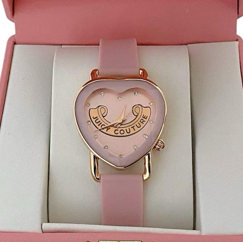 May include: A heart-shaped analog watch with a rose gold-tone case and a light pink silicone strap. The watch face features a light pink background, small crystal hour markers, and the text Juicy Couture printed in a gold-tone scroll design.
