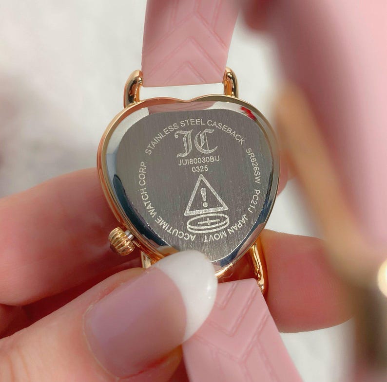 May include: A heart-shaped gold-tone watch case with a stainless steel back featuring the text STAINLESS STEEL CASEBACK, JC, JU180030BU, 0325, SR626SW, PC21J, JAPAN MOVT, ACCUTIME WATCH CORP, and a warning symbol. It has a light pink textured strap.
