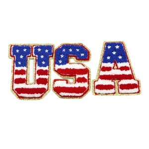 May include: USA spelled out in large, red, white, and blue letters with gold glitter trim. Each letter is decorated with a stylized American flag pattern.