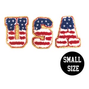 May include: Three felt letters spelling "USA" in a gold outline. Each letter is decorated with a red, white, and blue American flag pattern. The letters are small in size.