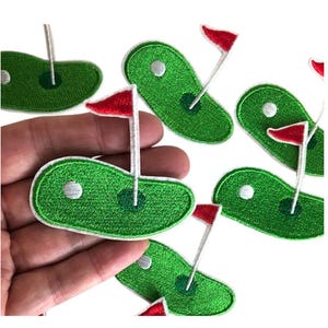 May include: A set of six green and white embroidered iron-on patches featuring a golf green with a flag and a golf ball.