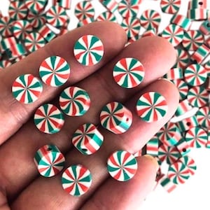 May include: A close-up of a hand holding a variety of red, white, and green candy cane-shaped beads. The beads are made of polymer clay and have a swirl design.