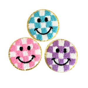 May include: Three round patches with a smiley face design. Each patch features a checkerboard pattern in blue, pink, and purple with white squares. The edges are trimmed with gold. The smiley faces have black eyes and a black curved mouth.