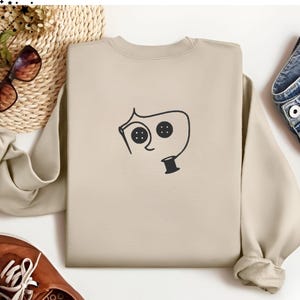 May include: A beige sweatshirt with a black graphic of a stylized face with buttons for eyes and a spool of thread for a mouth.