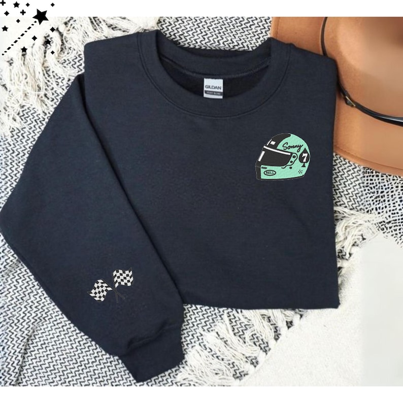 Op de afbeelding: Een marineblauw sweatshirt met een ronde hals en lange mouwen. De linkermouw heeft twee geborduurde geblokte vlaggen. De voorkant van het sweatshirt heeft een mintgroene racehelm-afbeelding met de naam "Sonny" en het nummer 7.