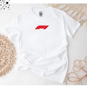 Op de afbeelding: Wit T-shirt met ronde hals en een rood F1-logo op de voorkant. Het logo is een gestileerd ontwerp met een rode balk en een schuine rode lijn. Het shirt is gemaakt van zacht, licht materiaal.