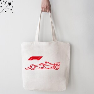 Op de afbeelding: Cr&egrave;mekleurige draagtas met een rode omtrek van een Formule 1-raceauto en het F1-logo. De tas heeft twee lange handvatten en wordt door een persoon omhooggehouden.