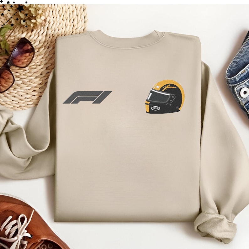 Op de afbeelding: Beige crewneck sweatshirt met een grijs F1-logo en een helm-afbeelding. De helm is zwart, geel en wit. De sweatshirt is gemaakt van een zacht materiaal. Het ontwerp staat op de borst.