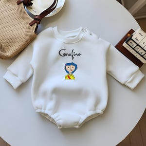 May include: White long-sleeved baby onesie with a cartoon image of Coraline from the movie Coraline. The onesie has a ruffled bottom and a snap closure at the crotch.
