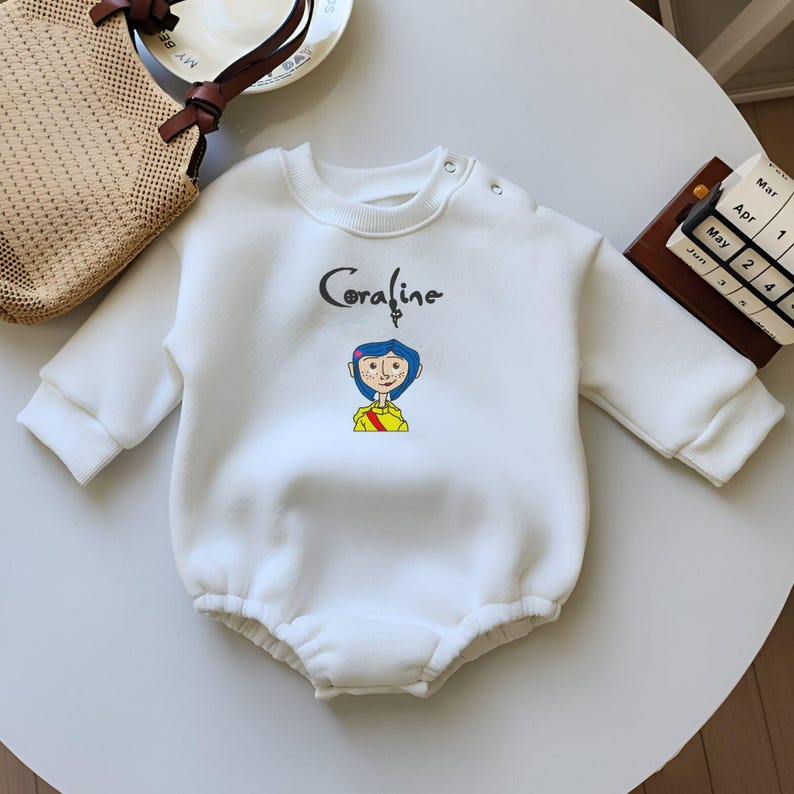 May include: White long-sleeved baby onesie with a cartoon image of Coraline from the movie Coraline. The onesie has a ruffled bottom and a snap closure at the crotch.