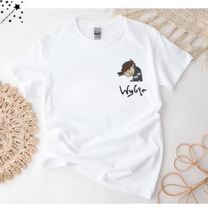 May include: White t-shirt with a cartoon illustration of a person with brown hair and a black and white outfit. The text "Wybie" is written below the illustration.