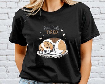 Op de afbeelding: Zwart T-shirt met een cartoon illustratie van een slapende beagle. De beagle is bruin en wit, liggend op een wit oppervlak. De tekst "Pawsitively TIRED" staat boven de hond, met pootafdrukken.