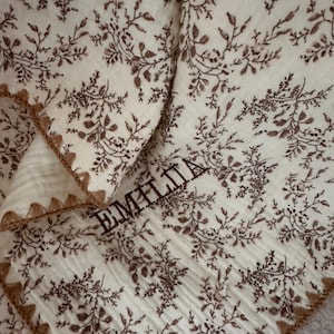 Op de afbeelding: Crèmekleurige stof met een bruin bloemenpatroon en de naam "EMILIA" in bruin geborduurd. De randen zijn afgewerkt met een bruine, gefranjerde rand. De stof is gevouwen en toont meerdere lagen.
