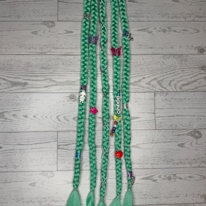 May include: Braided turquoise hair extensions with decorative butterfly, flower, and charm accents. The extensions are attached to a black hair tie. The word "DREAM" is spelled out with white beads on one braid. The braids have a textured, woven appearance.