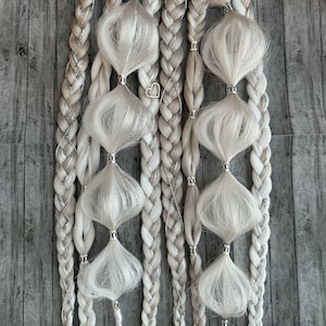 Può includere: Primo piano di due set di extension per capelli intrecciati bianchi e argentati. Ogni treccia presenta dettagli decorativi argentati e sezioni bianche soffici. Le trecce sono disposte verticalmente su uno sfondo di legno grigio.