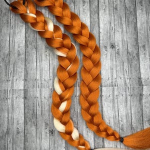 May include: Two orange braided hair extensions with black elastic bands at the top and bottom. Each braid has a section of white hair woven into the orange strands. The extensions are displayed on a gray wooden surface.