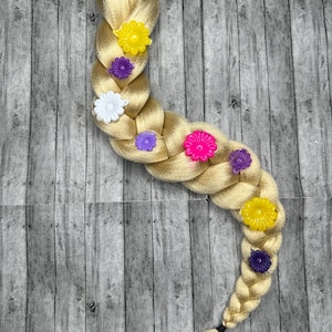 Pode incluir: Uma extensão de cabelo trançado loiro com uma faixa elástica preta no topo e uma extremidade texturizada. A trança é decorada com enfeites em forma de flor coloridos em amarelo, roxo, branco e rosa.