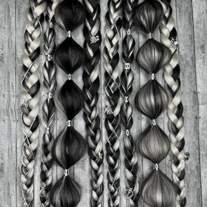 Puede incluir: Seis extensiones de cabello trenzadas en blanco y negro con cuentas y abalorios plateados. Las extensiones cuelgan sobre un fondo de veta de madera.