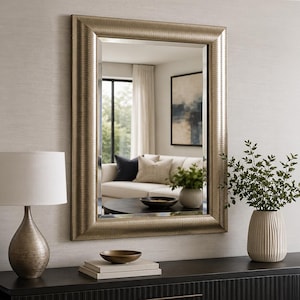 May include: A rectangular wall mirror featuring a wide, metallic champagne-coloured frame with a subtle horizontal textured finish. The mirror has bevelled edges and is mounted on a wall, reflecting a living room interior with a sofa and decor.