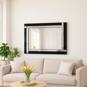 May include: A rectangular wall mirror featuring a wide, glossy black frame accented with thin, polished silver-toned metallic strips. The mirror has a clear, reflective glass surface that displays a reflection of a room with a white door and window.