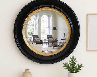 May include: A round wall mirror featuring a wide, matte black outer frame and a thinner, polished gold inner rim. The reflective glass surface shows a distorted, wide-angle view of a room with furniture, windows, and a floor lamp.