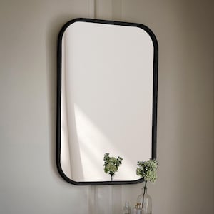 May include: A rectangular mirror with rounded corners, framed in black. The mirror is hanging on a white wall and reflects light from a window.