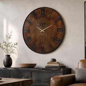 May include: A large circular wall clock with a dark, weathered bronze-toned face and a thin black outer rim. The clock features prominent, raised black Roman numerals and two slender, straight gold-coloured hands centred on the dial.