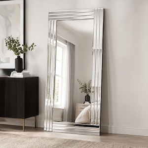 May include: A tall, rectangular floor mirror featuring a wide, multi-panelled frame with a reflective, mirrored finish. The frame consists of several vertical and horizontal strips of glass that create a layered, geometric border around the centre.